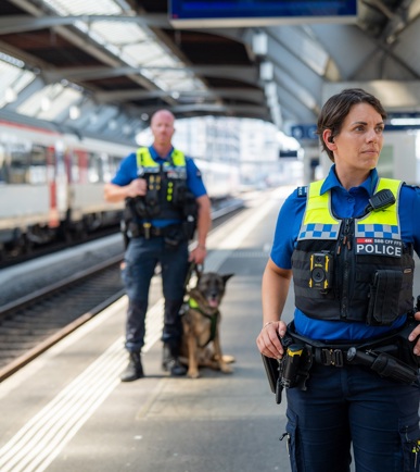 Zwei SBB Beamte stehen auf dem Perron in einem Bahnhof. Einer der Beamten ist näher an der Kamera und ist weiblich, der hintere ist männlich und hat einen Deutschenschäferhund neben sich